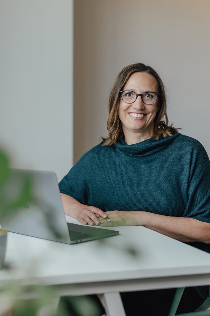 Coachin sitzt lächelnd vor ihrem Laptop am Schreibtisch.